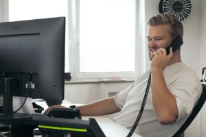 Junger Mann sitzt an einem Schreibtisch mit Computer. In seiner Hand hat er den Hörer eines Festnetztelefons.