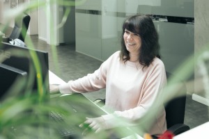Dunkelhaarige Frau sitzt lachend an einem Schreibtisch mit Computer.