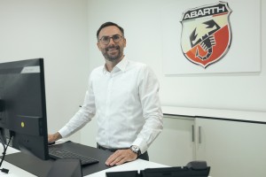 Mann in weißem Hemd steht an seinem hochgefahrenen Schreibtisch. Hinter ihm an der Wand das Logo der Automarke Abarth.