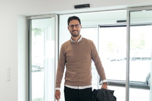 Mann mit Wollpulli, Chinohose und weißen Sneakern kommt lächelnd zur Schiebetür der Augustin Group hinein.