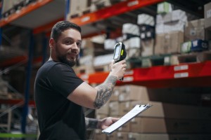 Junger Mann mit Bart und tätowiertem Unterarm steht mit Scanner vor einem Hochregal.